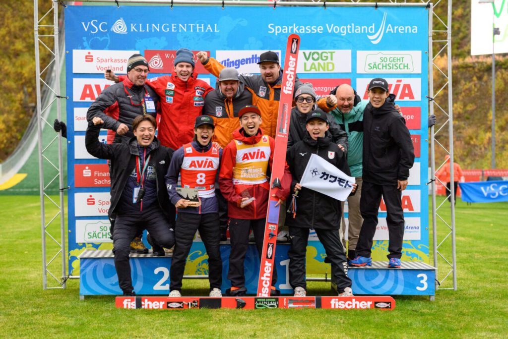 Das japanische Team jubelt auf dem Siegerpodest in der Sparkasse Vogtland Arena in Klingenthal, in der Mitte ein Springer im gelben Trikot des Grand-Prix-Gesamtführenden.
