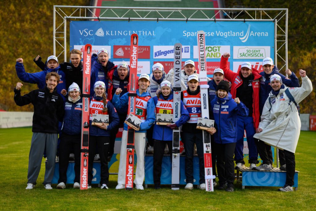 Siegerehrung des Mixed-Team-Wettbewerbs in der Sparkasse Vogtland Arena in Klingenthal, die Teams jubeln vor der Sponsorenwand.